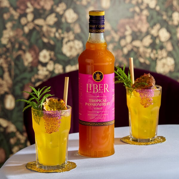 A bottle of Liber & Co Tropical Passionfruit Syrup with two glasses of yellow tropical drinks garnished with passionfruit and herbs.