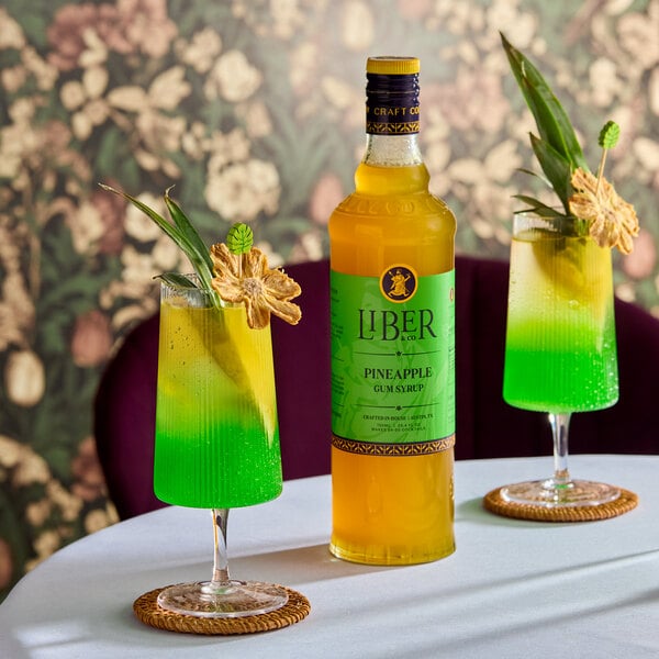 A bottle of Liber & Co Pineapple Gum Syrup 750 mL is displayed on a table with two green and yellow cocktails garnished with pineapple and leaves.