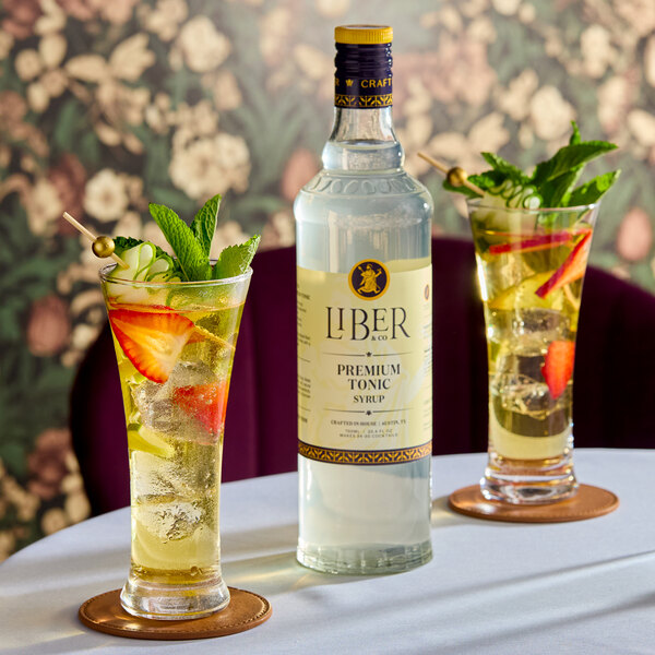 A bottle of Liber & Co Premium Tonic Syrup 750 mL placed on a table with two garnished glasses of tonic drinks.