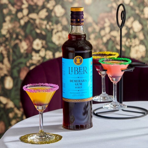 A 750 mL bottle of Liber & Co Demerara Gum Syrup placed on a table with colorful cocktails.
