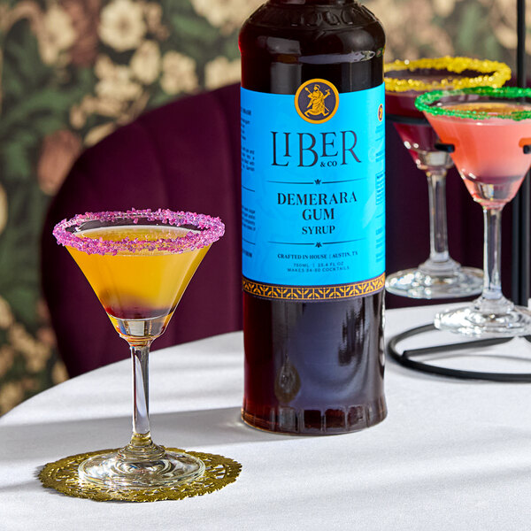 A 750 mL bottle of Liber & Co Demerara Gum Syrup displayed next to colorful cocktails on a table.