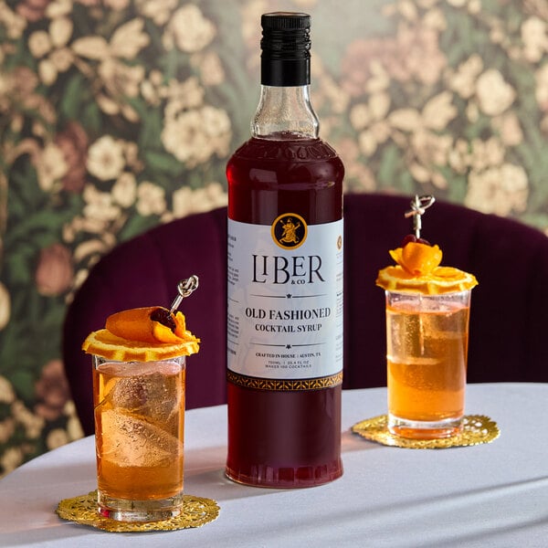 A 750 mL bottle of Liber & Co Old Fashioned Cocktail Syrup placed on a table with two garnished cocktails.