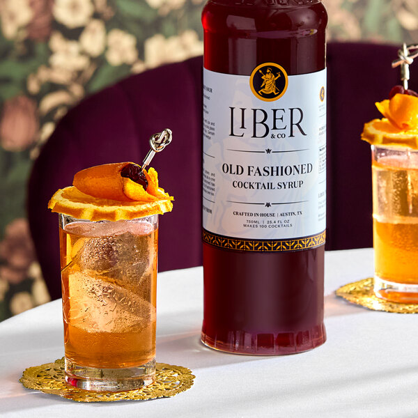A bottle of Liber & Co Old Fashioned Cocktail Syrup, 750 mL, displayed on a table with garnished cocktails.
