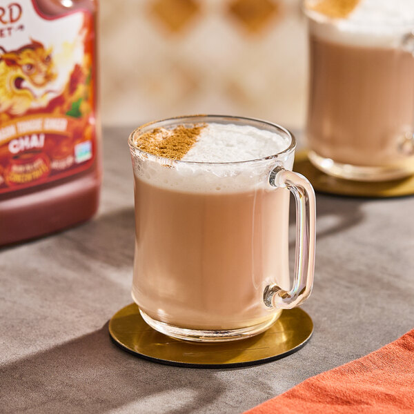 A glass mug filled with a chai tea latte topped with foam and a sprinkle of spice, placed on a coaster.
