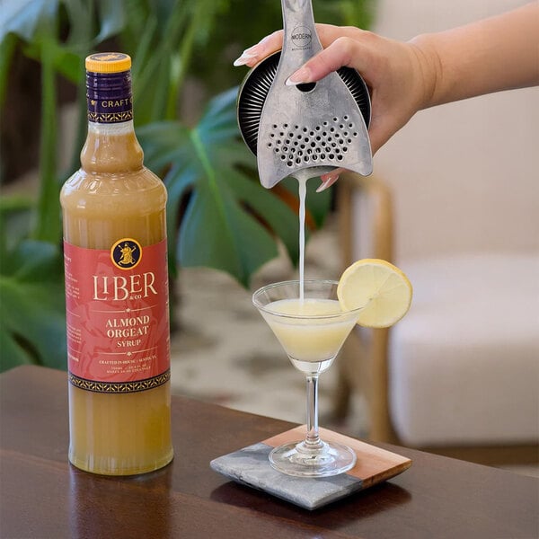 A bottle of Liber & Co Almond Orgeat Syrup next to a cocktail being strained into a glass with a lemon garnish.