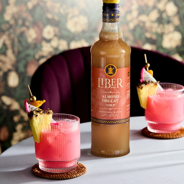 A 750 mL bottle of Liber & Co Almond Orgeat Syrup placed on a table with two pink cocktails garnished with fruit.