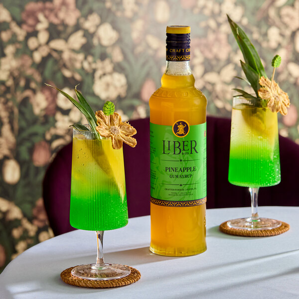 A bottle of Liber & Co Pineapple Gum Syrup 750 mL is displayed on a table with two green and yellow cocktails garnished with pineapple and leaves.