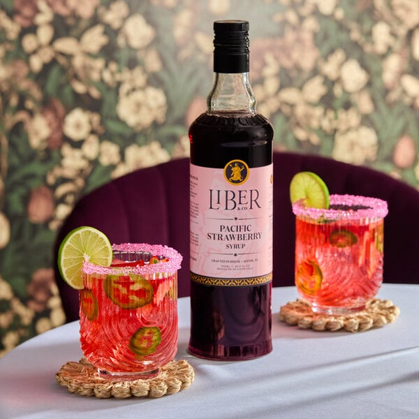 A bottle of Liber & Co Pacific Strawberry Syrup with two glasses of a pink beverage garnished with lime slices and pink sugar rims.