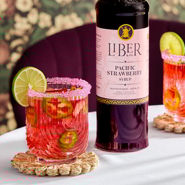 A bottle of Liber & Co Pacific Strawberry Syrup next to a glass of pink cocktail garnished with lime and jalapeño slices.