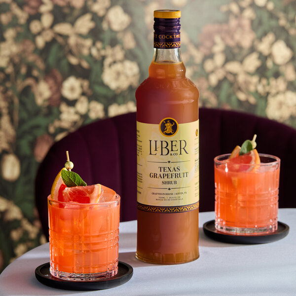 A bottle of Liber & Co Texas Grapefruit Shrub 750 mL placed on a table with two grapefruit cocktails.