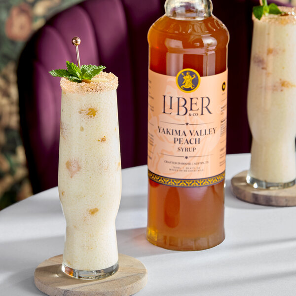 A bottle of Liber & Co Yakima Valley Peach Syrup (750 mL) placed next to a tall glass of a creamy peach beverage garnished with mint.