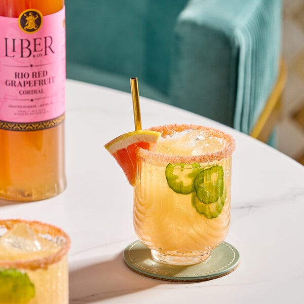 A bottle of Liber & Co Rio Red Grapefruit Cordial Syrup next to a cocktail garnished with grapefruit, jalapeño slices, and a sugared rim.