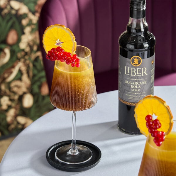 A bottle of Liber & Co Sugarcane Kola Syrup next to a cocktail garnished with an orange slice and red berries.