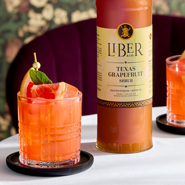 A bottle of Liber & Co Texas Grapefruit Shrub and a glass of grapefruit cocktail garnished with a grapefruit wedge and mint.