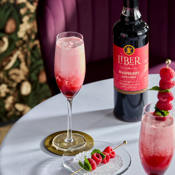 A bottle of Liber & Co Raspberry Gum Syrup alongside a raspberry cocktail and fresh raspberries on a table.