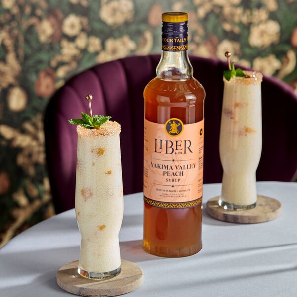 A 750 mL bottle of Liber & Co Yakima Valley Peach Syrup displayed on a table with two tall peach-flavored drinks garnished with mint and sugar rims.