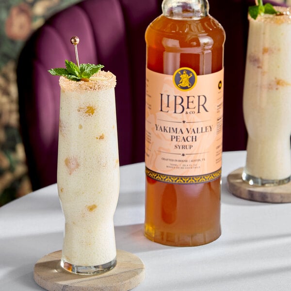 A bottle of Liber & Co Yakima Valley Peach Syrup next to a tall glass of a peach-flavored beverage garnished with mint.