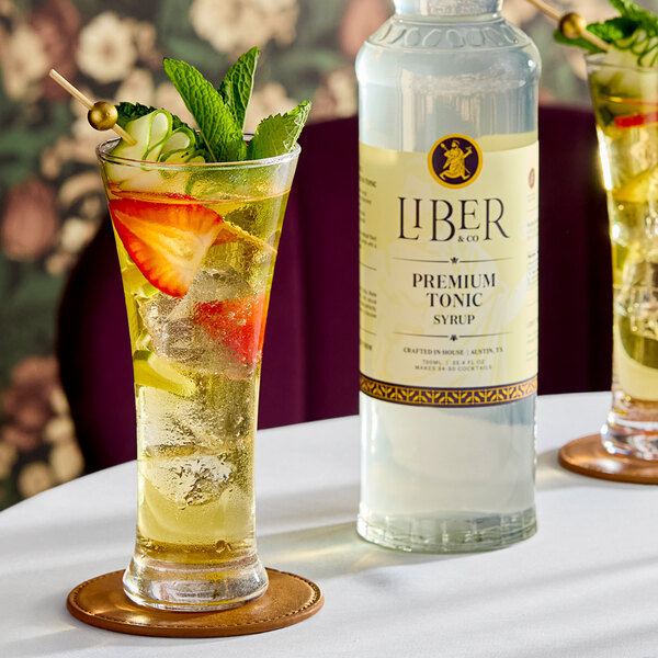 A bottle of Liber & Co Premium Tonic Syrup next to a glass of tonic-based cocktail garnished with fruit and mint.