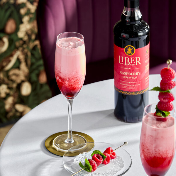 A bottle of Liber & Co Raspberry Gum Syrup 750 mL next to a glass of a pink raspberry beverage and a plate with fresh raspberries and mint.
