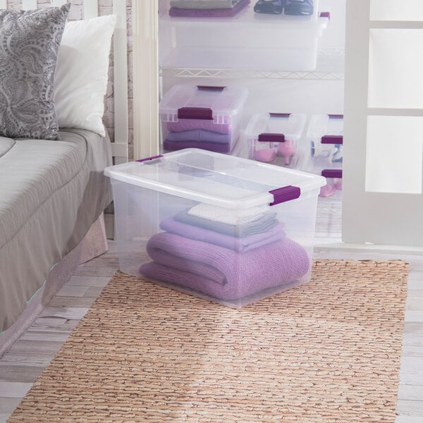 A clear plastic storage box with a latching lid and purple handles, partially filled with folded towels.