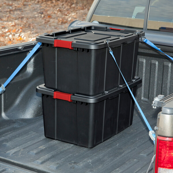 A pair of black industrial storage totes with red handle lids secured in the bed of a pickup truck.
