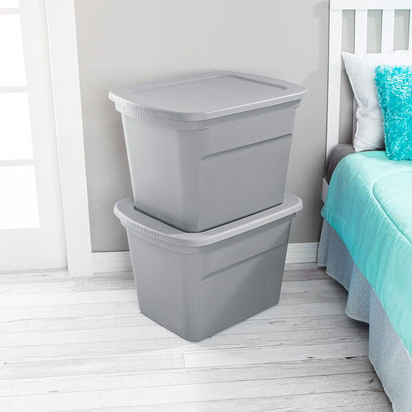 A pair of gray plastic storage totes with lids stacked next to a bed.