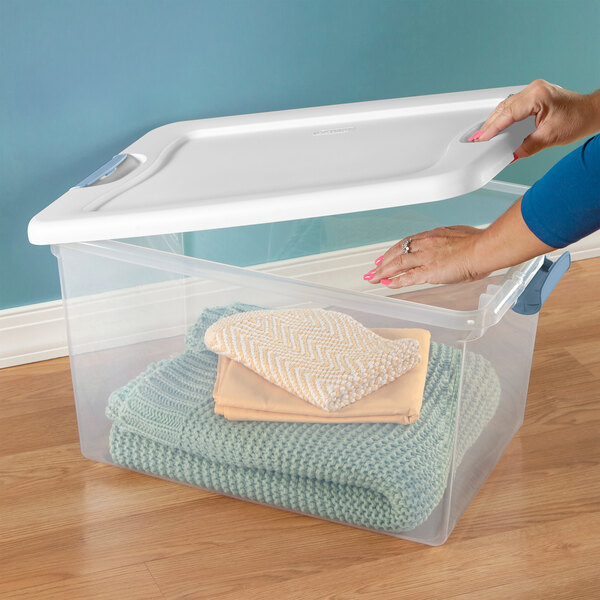 A clear plastic storage box with a white lid and blue handles, containing folded blankets and towels.
