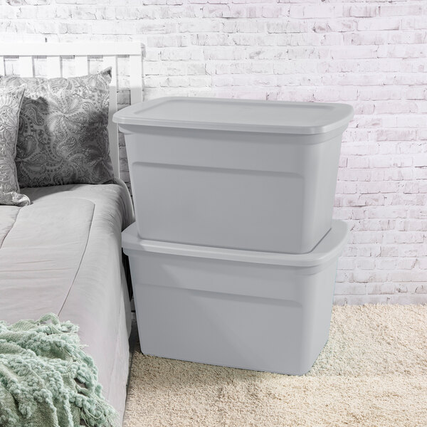A pair of large gray plastic storage totes with lids stacked next to a bed.