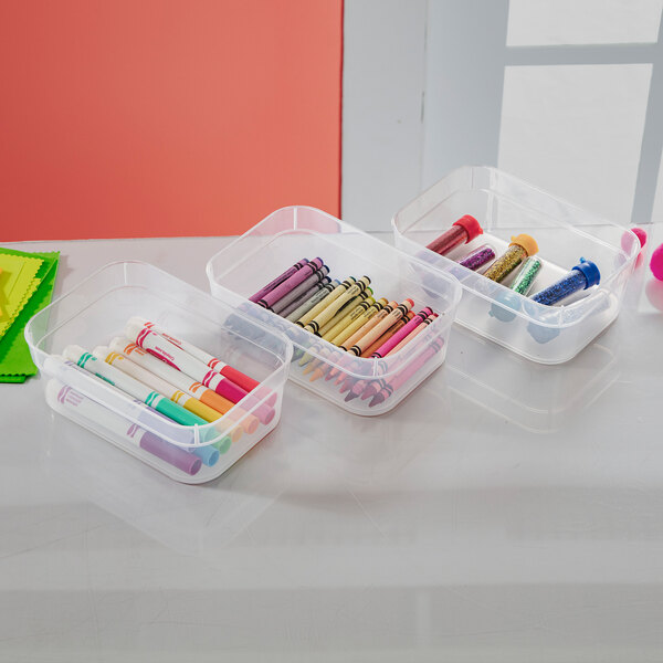 A set of clear, rectangular stackable storage trays containing art supplies such as markers, crayons, and glitter tubes.