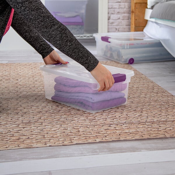 A clear plastic storage box with a white lid and purple handles, containing folded purple clothing.