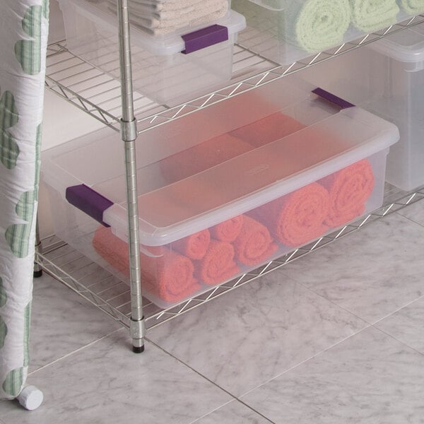 A clear plastic storage box with a purple handle lid, shown on a wire shelf and filled with orange towels.