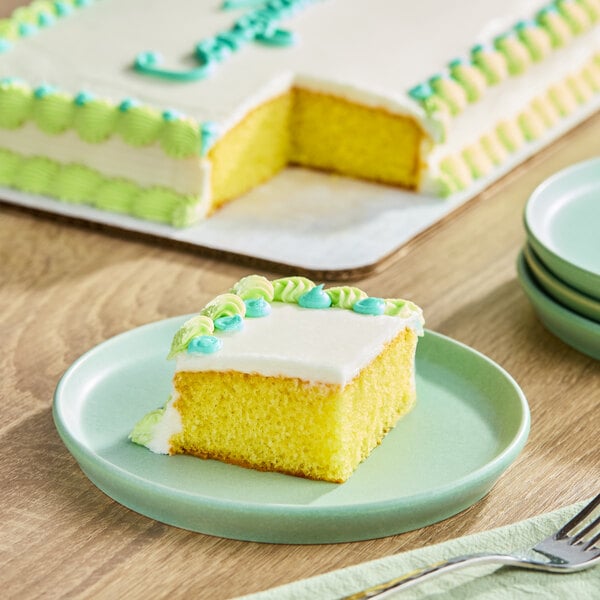 A slice of yellow sheet cake with white buttercreme icing and green and blue decorative frosting on a plate, with the remaining cake in the background.