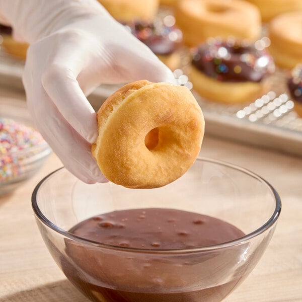 A gloved hand holding a plain mini yeast donut ring above a bowl of chocolate glaze, with more donuts in the background.