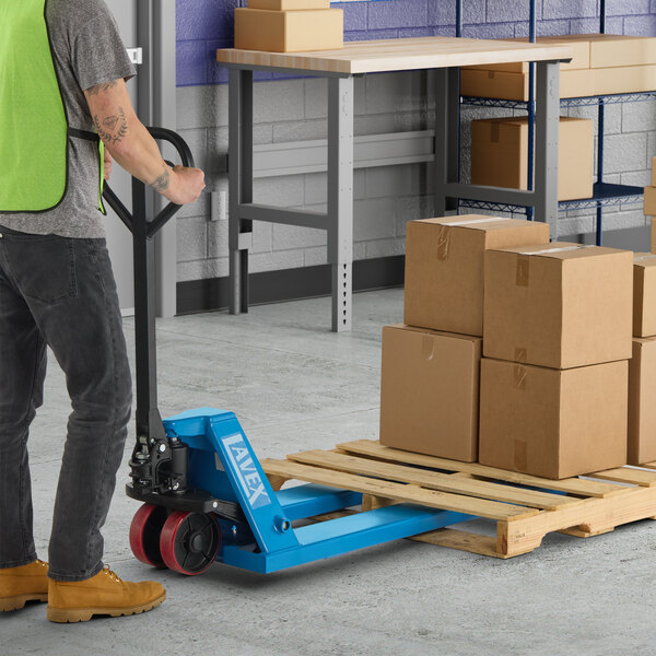 A blue manual pallet jack with 21-inch by 48-inch forks being used to move a wooden pallet loaded with cardboard boxes in a warehouse setting.