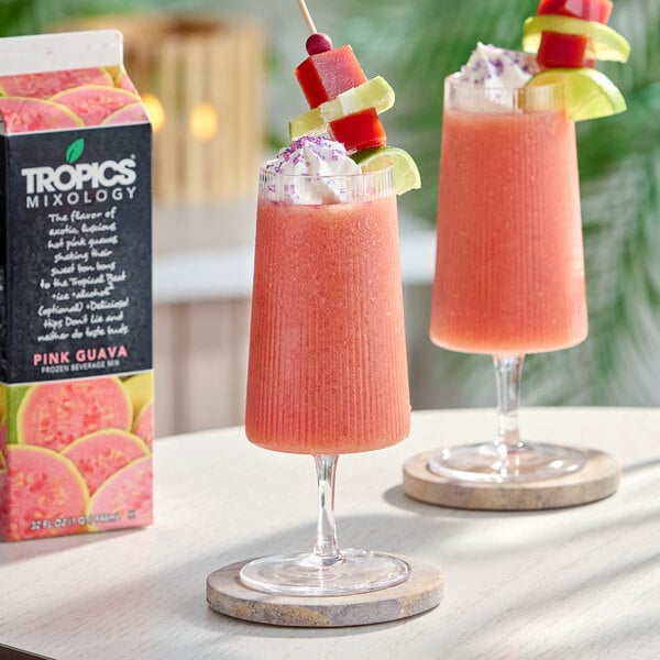 A carton of Tropics Mixology Pink Guava Frozen Beverage Mix next to two glasses of pink guava frozen drinks garnished with fruit and whipped topping.