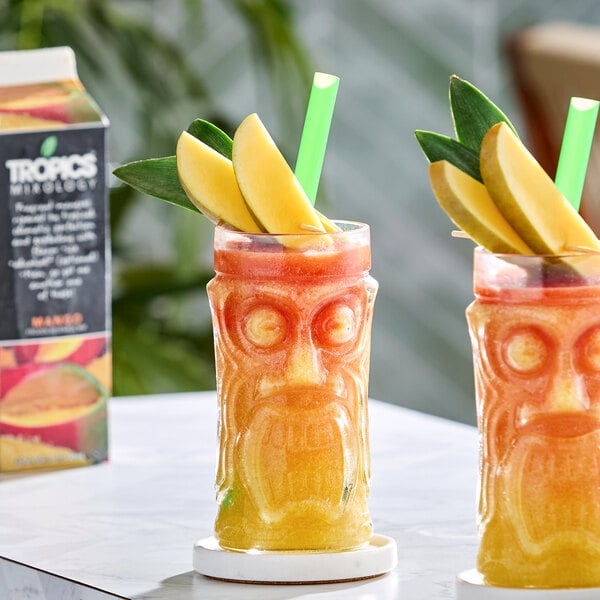 A glass of mango frozen beverage garnished with mango slices and a green straw, with a carton of Tropics Mixology Mango Frozen Beverage Mix in the background.