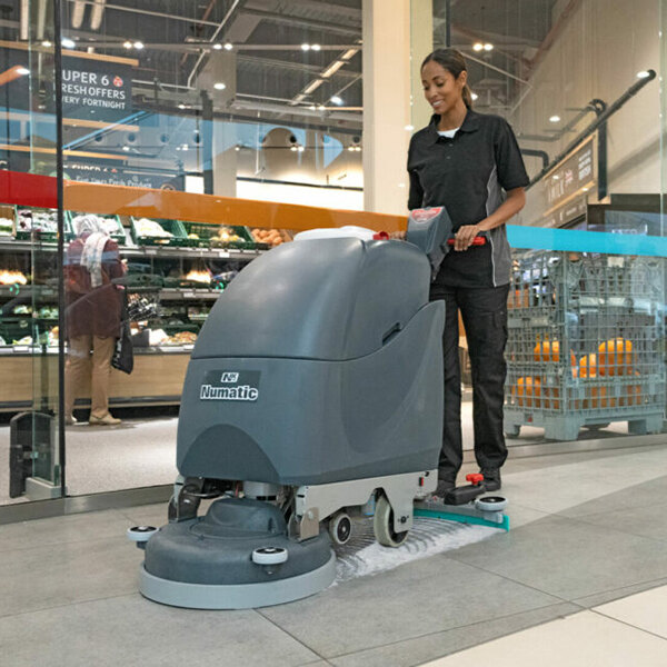 A Numatic Nacecare Solutions TBL 1120 20-inch lithium-ion walk-behind disc floor scrubber being used to clean a tiled floor in a commercial setting.