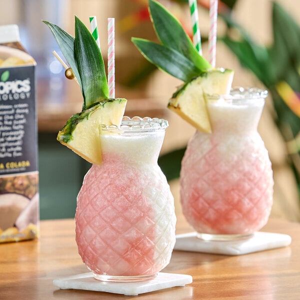 A pair of pink and white frozen pina colada drinks served in pineapple-shaped glasses, garnished with pineapple slices and leaves.