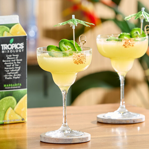 A carton of Tropics Mixology Margarita Frozen Beverage Mix next to two margarita glasses garnished with jalapeño slices and cocktail umbrellas.