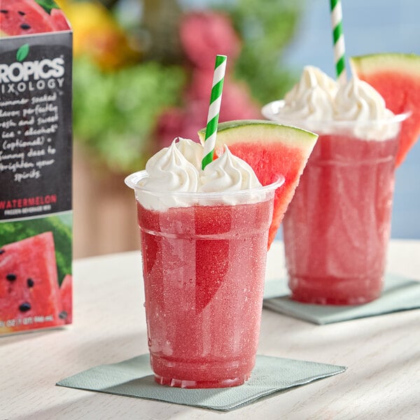 A plastic cup filled with a pink frozen watermelon beverage, topped with whipped cream, a green-striped straw, and a slice of watermelon, sitting on a napkin.