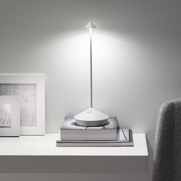 A modern white cordless rechargeable table lamp with a slim stem and conical shade, placed on a stack of books on a white shelf.
