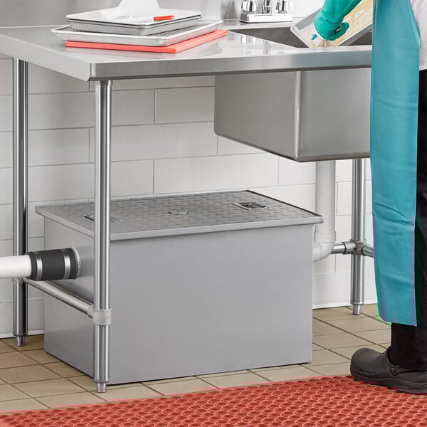 A rectangular metal grease trap with a diamond-patterned lid installed under a commercial kitchen sink.