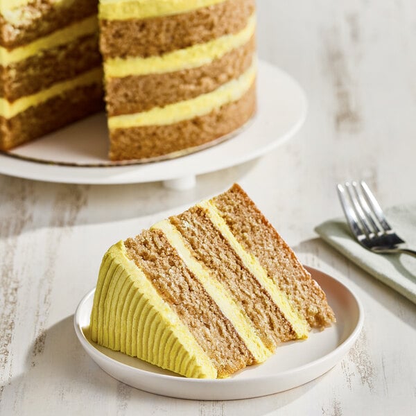 A slice of layered vegan lemon cake with yellow frosting on a white plate, with the rest of the cake in the background.