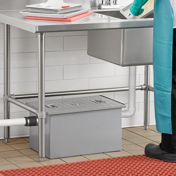 A rectangular gray grease trap with a metal lid and two non-threaded pipe connections, installed under a commercial kitchen sink.