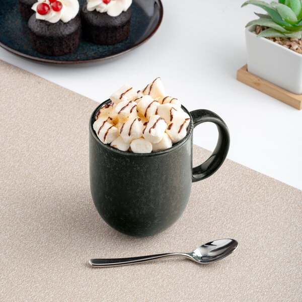A black porcelain coupe mug filled with marshmallows and chocolate drizzle, placed on a beige mat with a spoon nearby.