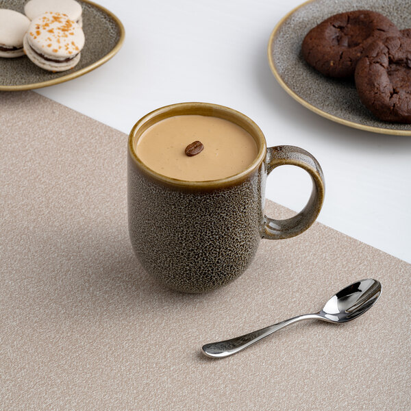 A brown porcelain coupe mug filled with a creamy beverage, placed on a beige placemat with a spoon nearby.