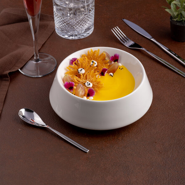 A white round porcelain coupe bowl filled with a yellow dessert and garnished with flowers and small decorative elements.