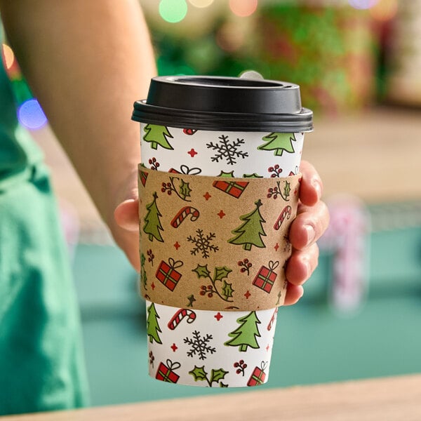 A disposable coffee cup with a festive Christmas-themed sleeve featuring cartoon trees, candy canes, presents, and holly.