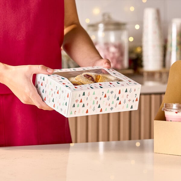 A rectangular bakery box with a modern Christmas print and a clear window on top, being held by a person.