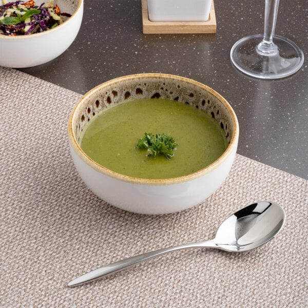 A round porcelain coupe bowl with a brown and cream spotted rim, filled with green soup and garnished with herbs.